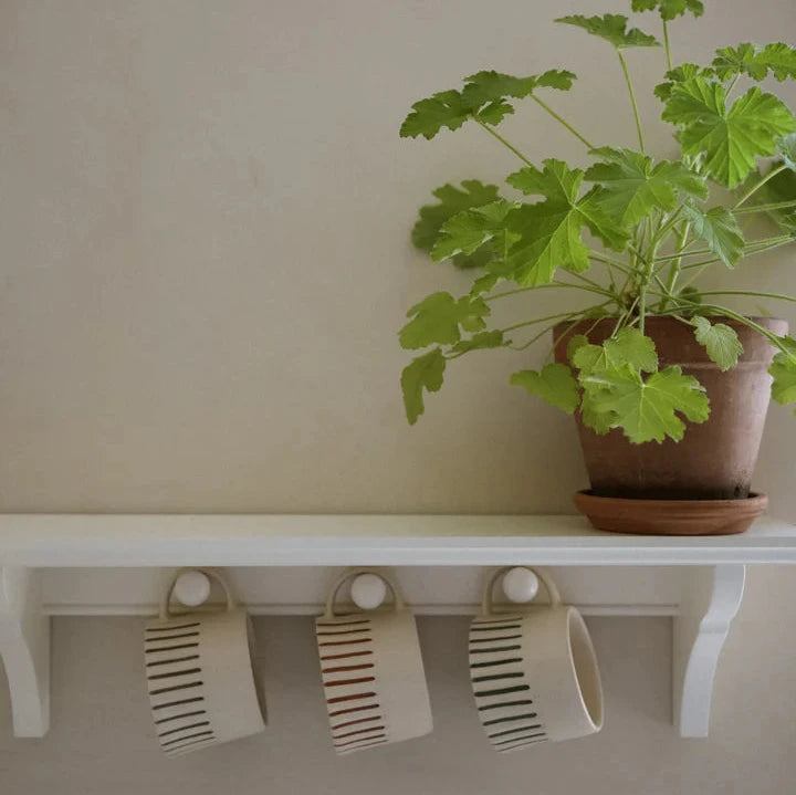 3 off white handmade mugs with rust coloured hand painted stripes, hanging from their handle on a white shelf with hooks.