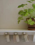 3 off white handmade mugs with rust coloured hand painted stripes, hanging from their handle on a white shelf with hooks.