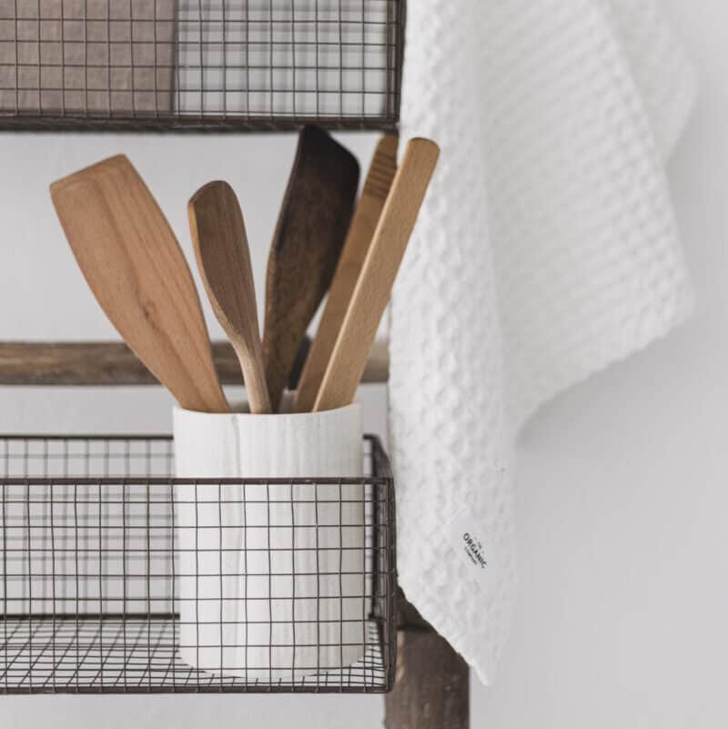 Close up of wire baskets on wooden storage ladder with kitchen utensils in white pot.