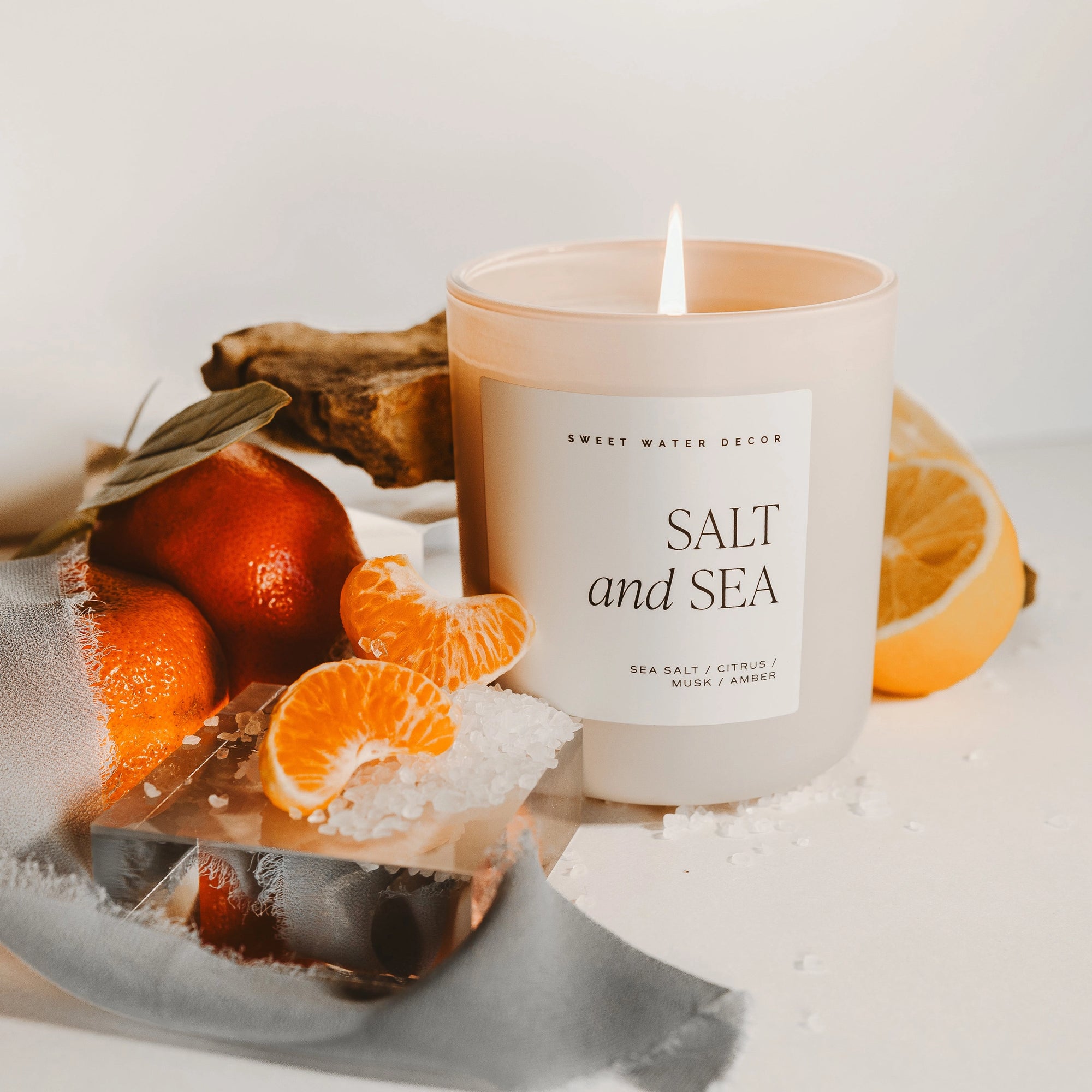 Close up salt and sea large soy candle in matte white jar, lit with oranges and salt.