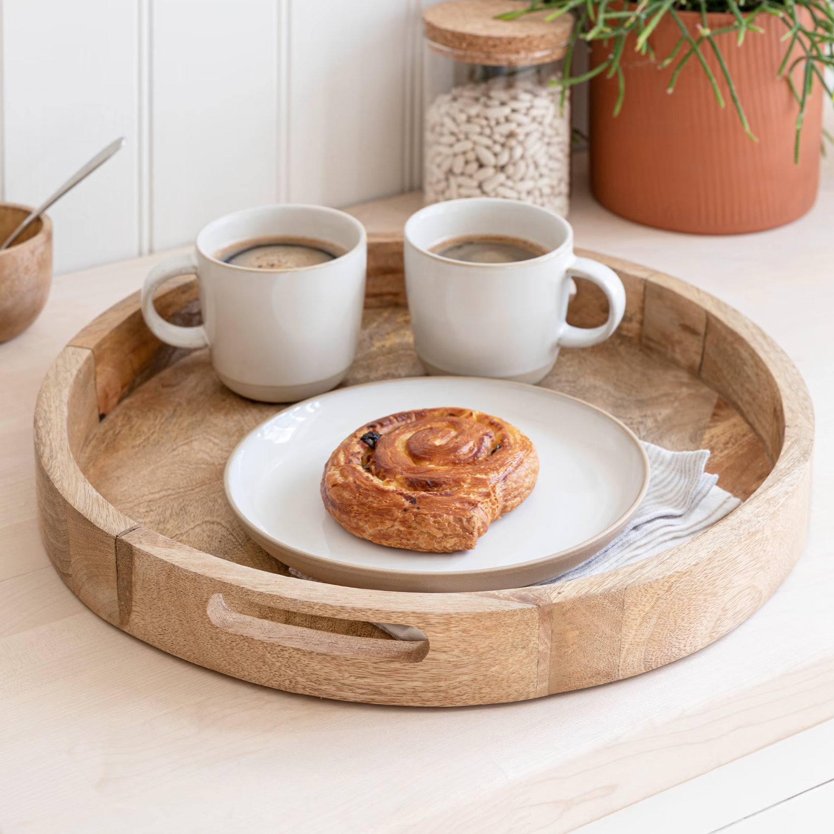 Round wooden tray with two cream coffee mugs and a pastry.