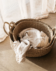 The set of two audrey seagrass baskets with a frilly linen towel draped inside. Flowing white curtain in background.