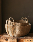 The Audrey Oval Seagrass Baskets - Set of Two placed on rustic wooden table against chocolate brown wall.