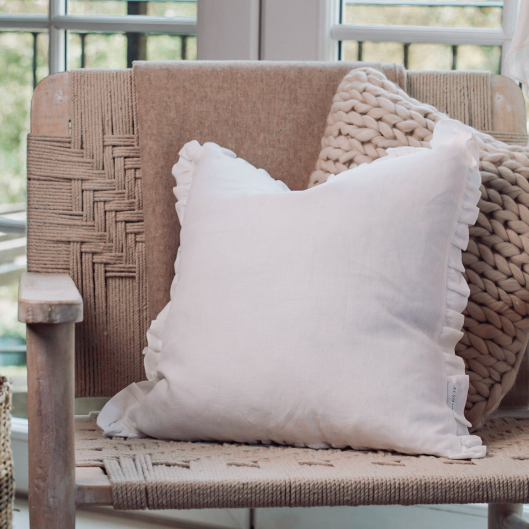 Lucie Linen White Ruffle Cushion on a woven armchair with a knitted cushion sitting behind.