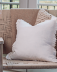 Lucie Linen White Ruffle Cushion on a woven armchair with a knitted cushion sitting behind.