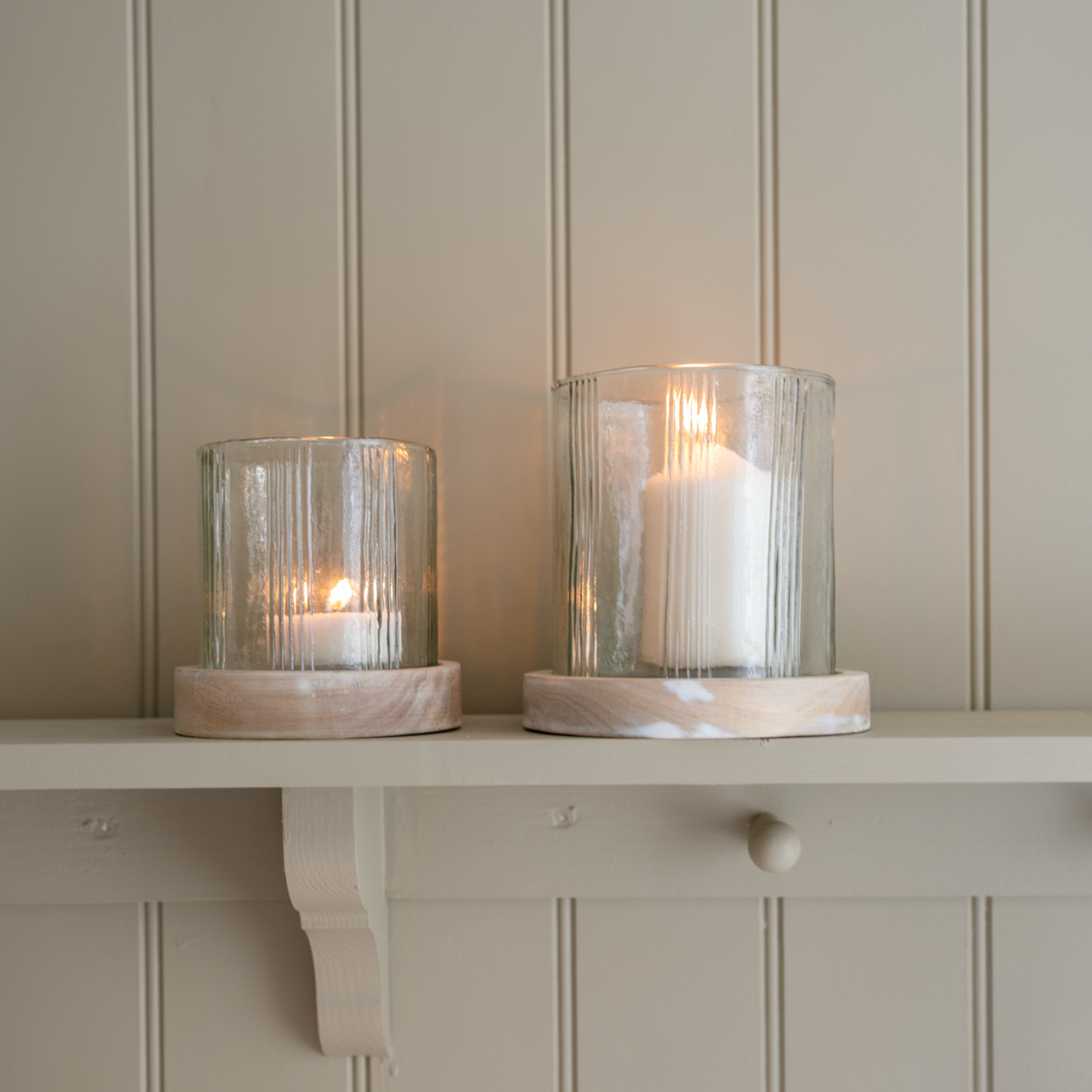 A set of two rustic glass candle holder against a panelled wall.