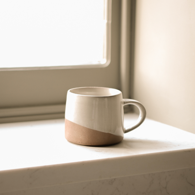 A loane stoneware mug with a slanted glaze sitting on a windowsil.