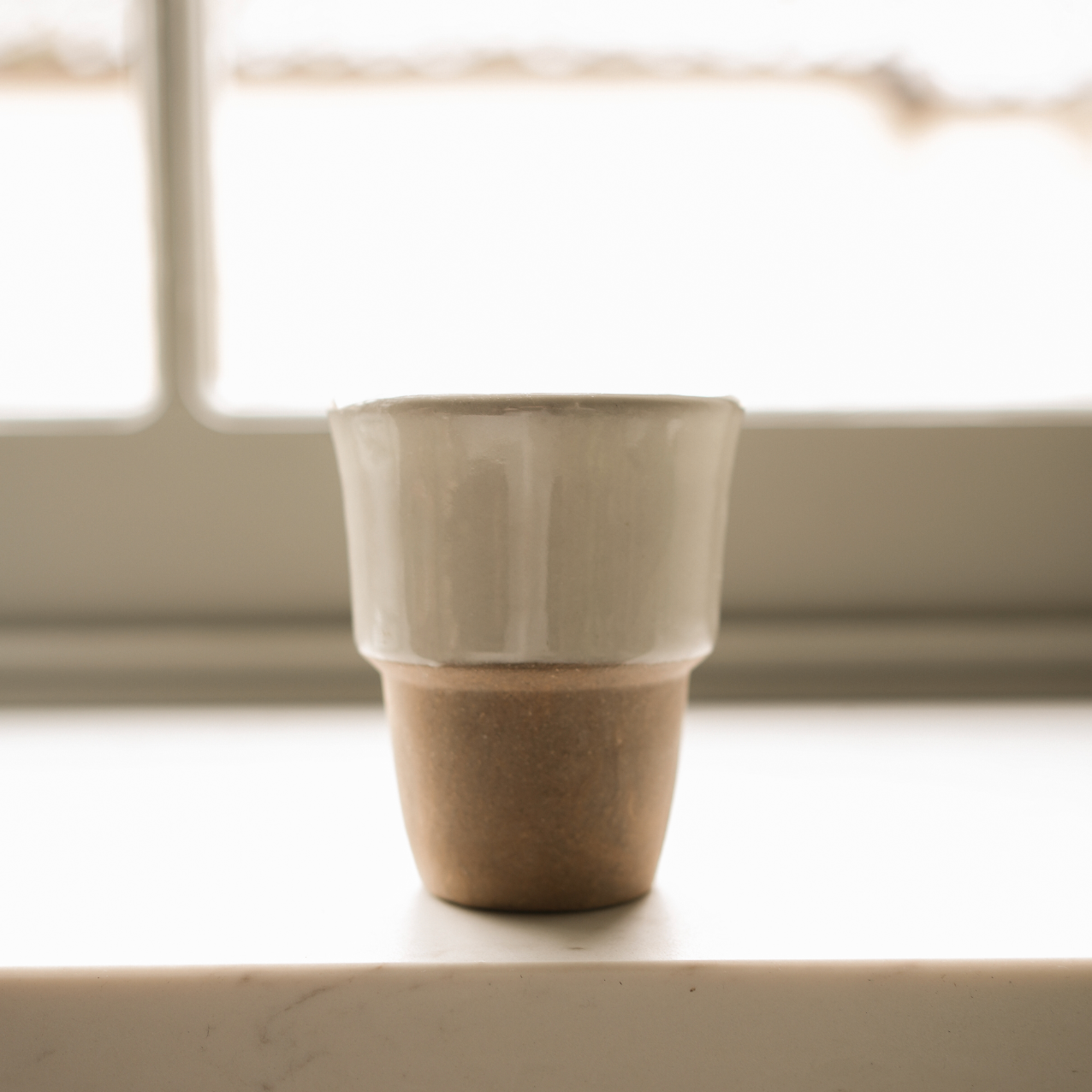 Stoneware Coffee Cup with a terracotta base and cream top in front of a window.