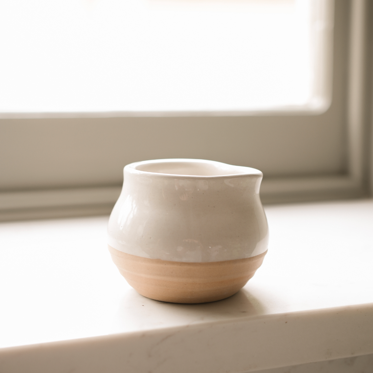 A lone bulbous stoneware milk jug sits in front of a window.