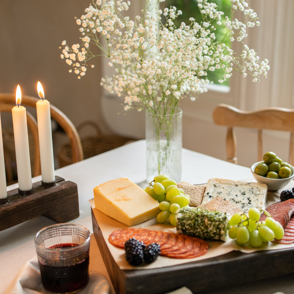 A dinner table with a cheese and fruit plater, flowers and some lit candles.