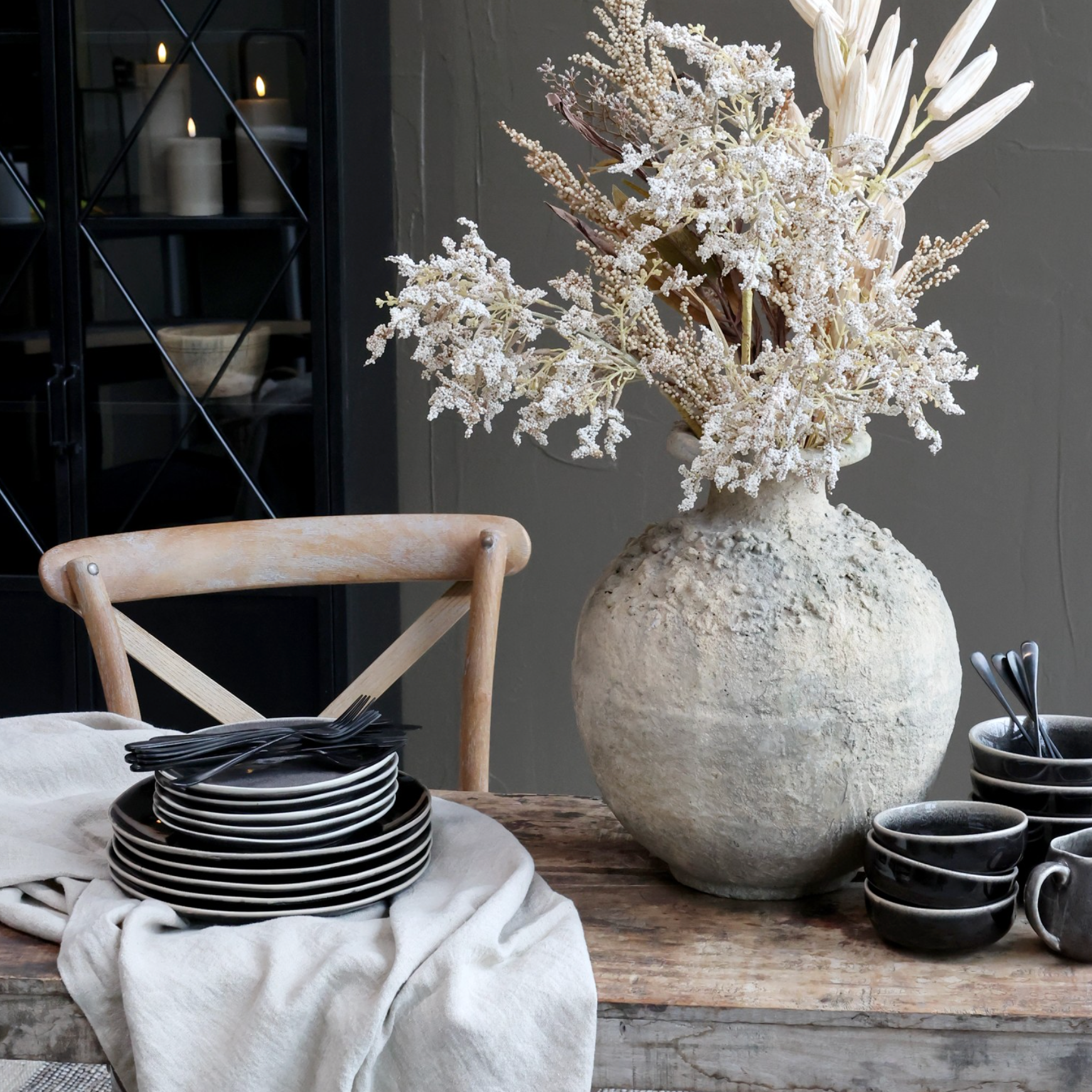 a dining table with a rustic vase with dried flowers, black crockery and a wooden dining chair.