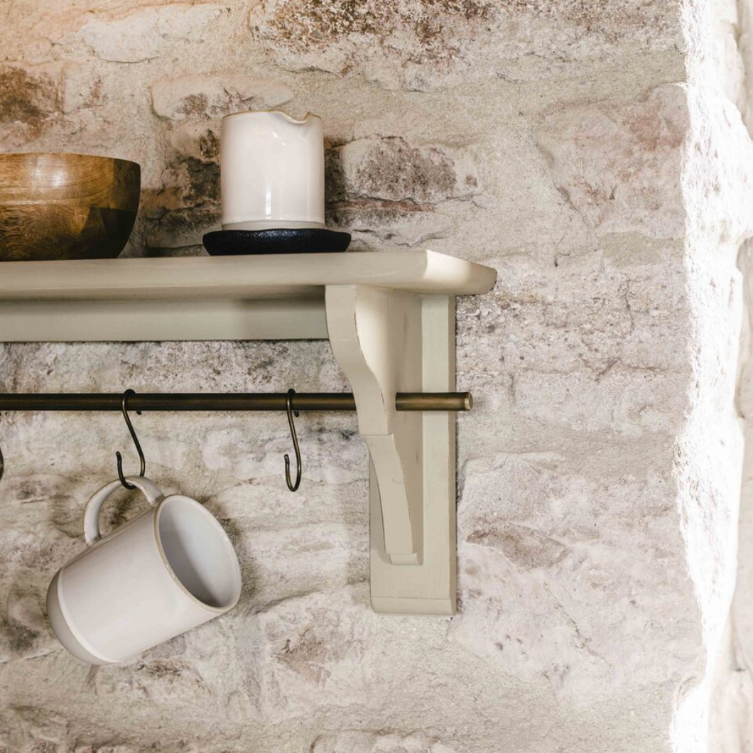 Clay coloured wooden shelf with brass shelf and hooks, attached to white stone wall with accessories.