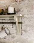 Clay coloured wooden shelf with brass shelf and hooks, attached to white stone wall with accessories.