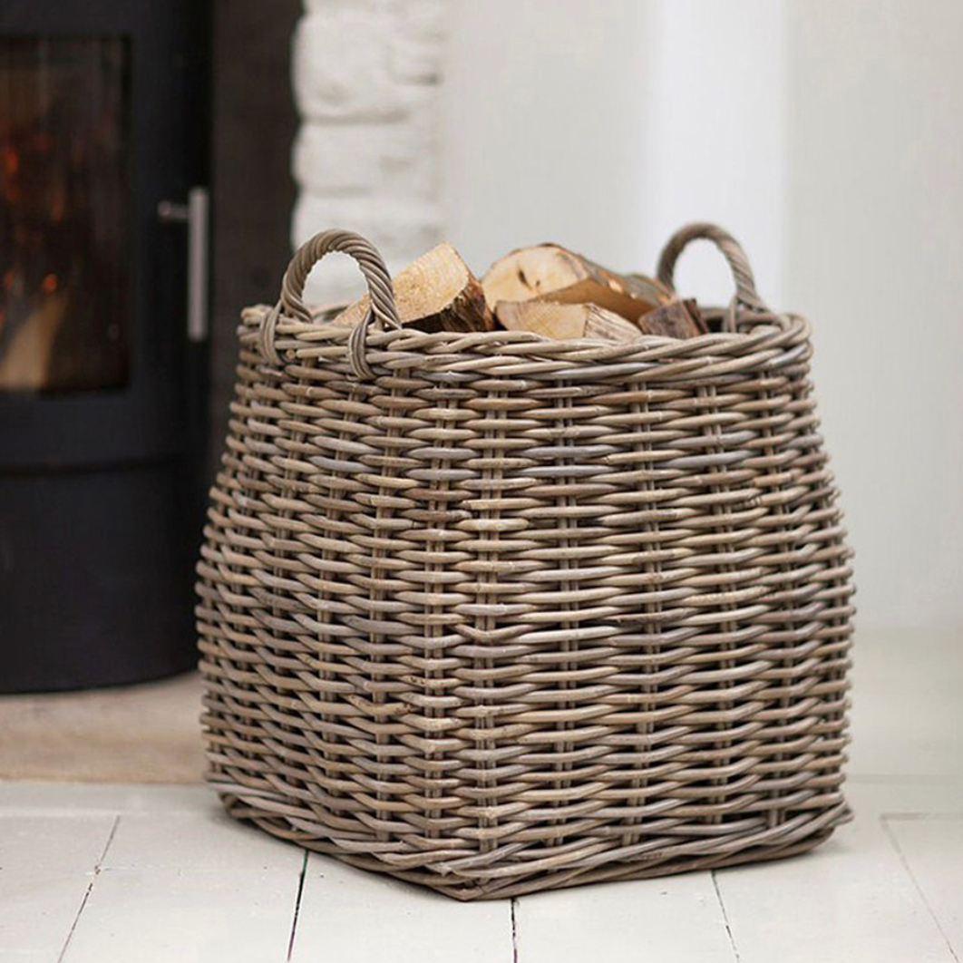 Tapered rattan log basket filled with logs in front of fire.