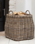 Tapered rattan log basket filled with logs in front of fire.