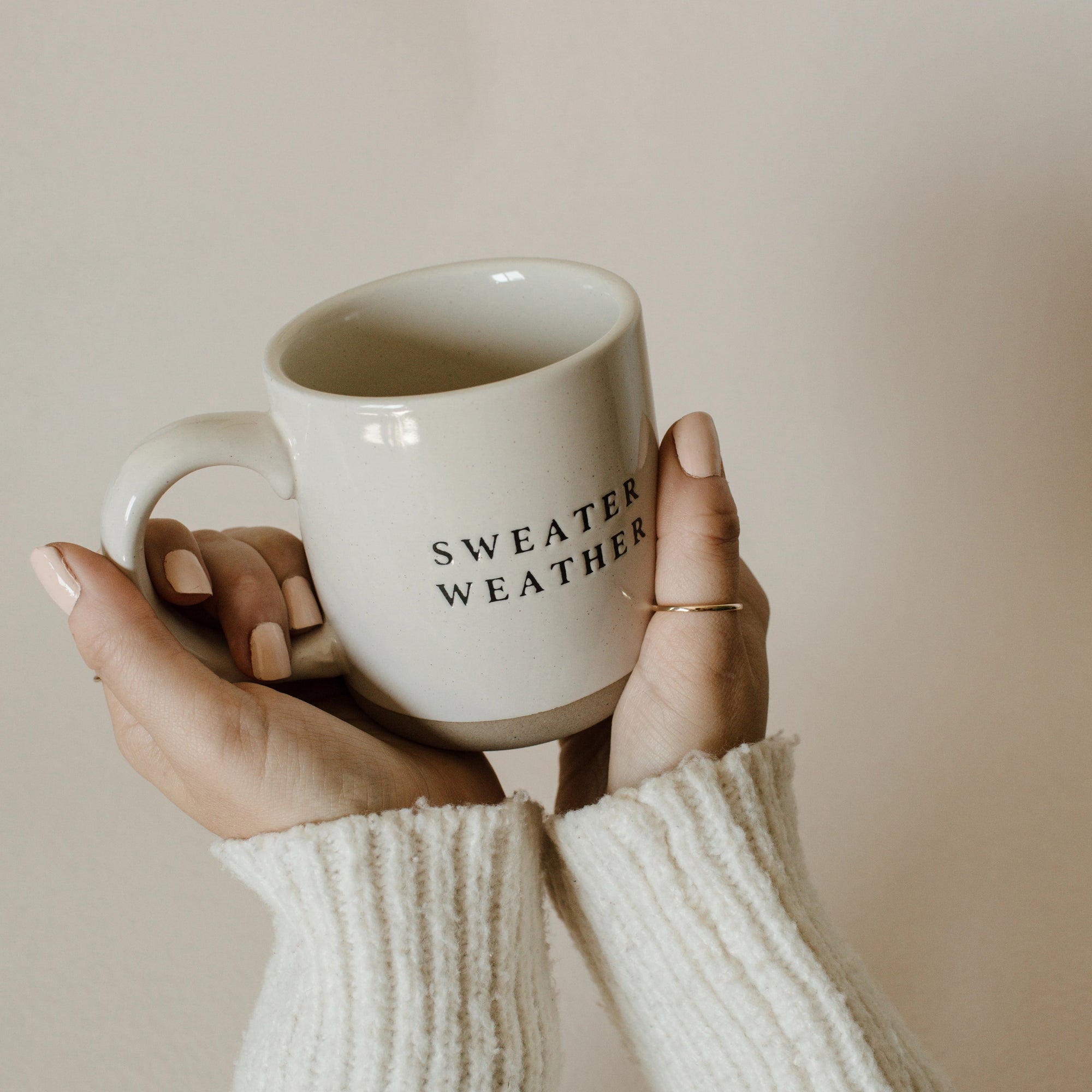 White and brown natural stoneware mug with black 'sweater weather' text, held in two hands.