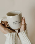 White and brown natural stoneware mug with black 'sweater weather' text, held in two hands.