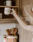 White and brown natural stoneware mug with black 'sweater weather' text, being put into a wooden kitchen cupboard with other mugs.