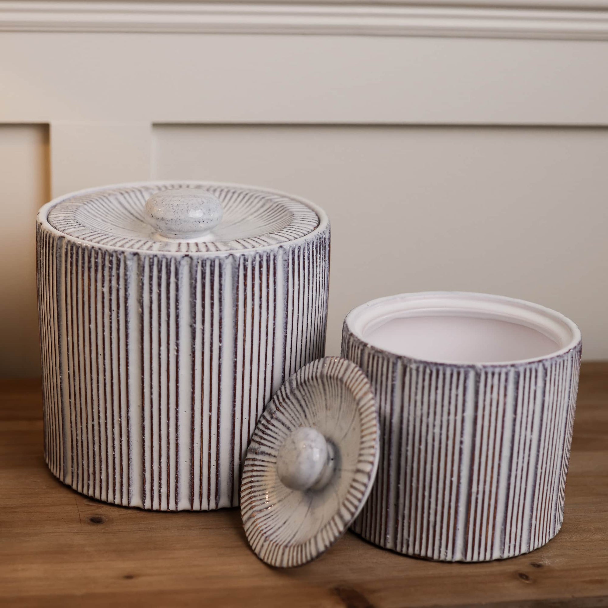 Decorative ceramic storage jars with red and white striped ridge and lid with small handle, on wooden console with lid off on smaller jar.