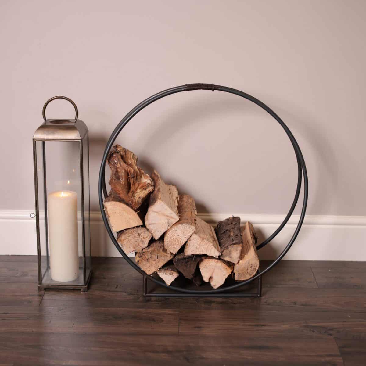Circular iron log carrier, filled with firewood, aside a candle lantern on a wood floor.