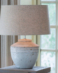 Medium white distressed lamp with neutral lamp shade on table in front of window.