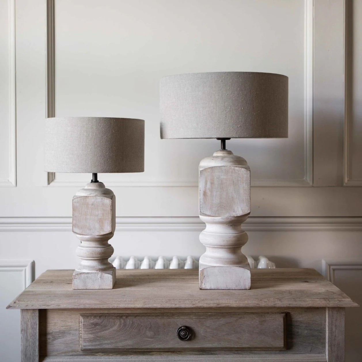 Large and small white washed mango wood table lamps on a wooden desk against a panelled wall. 