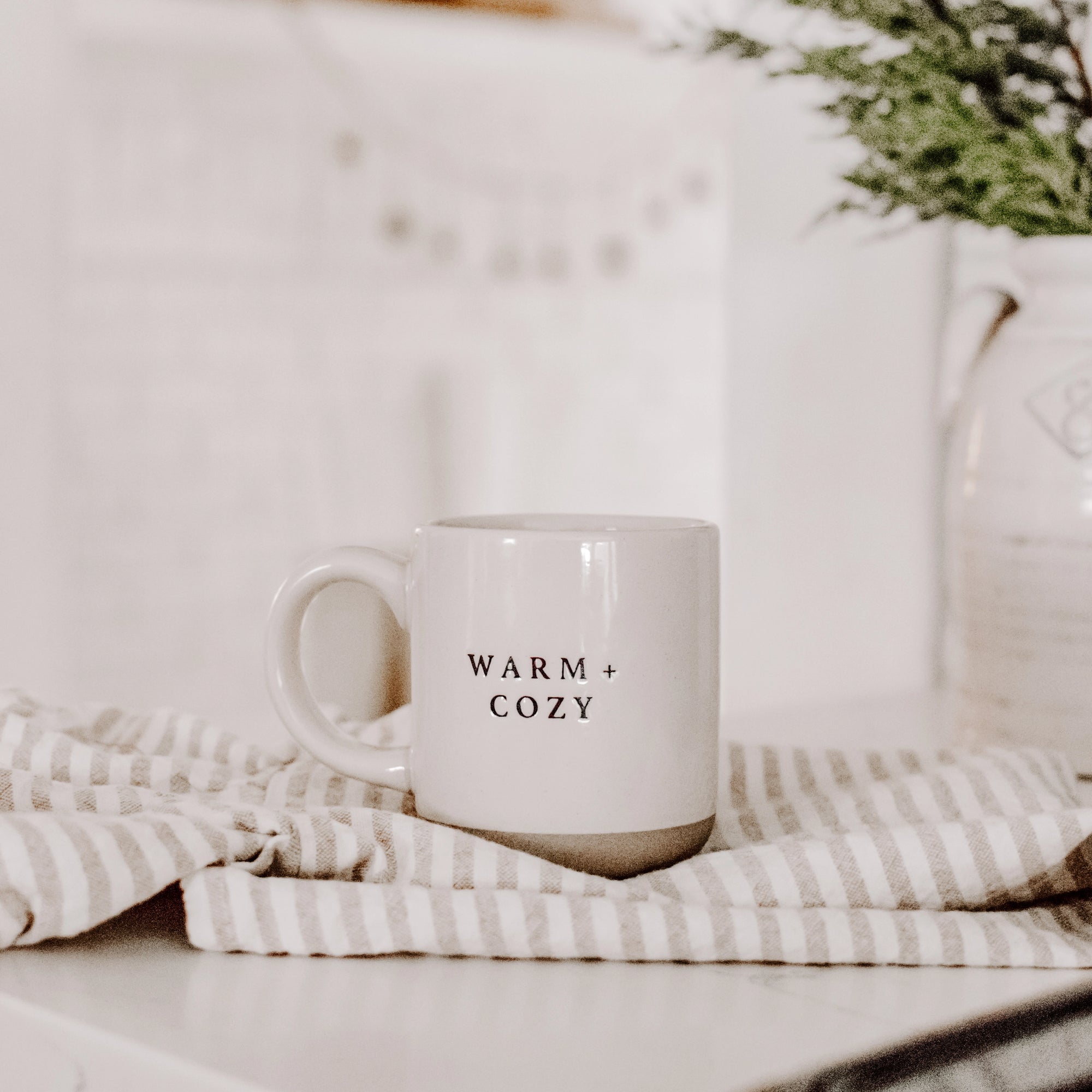 Cream and brown stoneware mug with black text 'warm + cozy' on the front, on striped tea towel.