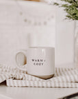 Cream and brown stoneware mug with black text 'warm + cozy' on the front, on striped tea towel.