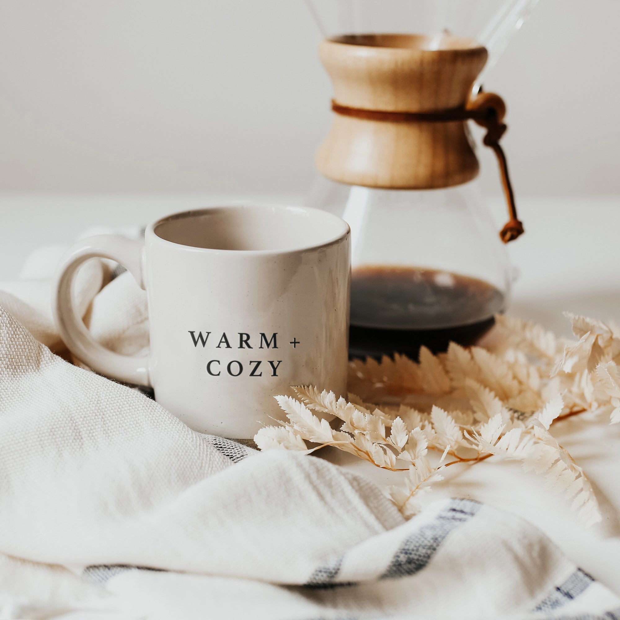 Cream and brown stoneware mug with black text 'warm + cozy' on the front, in front of coffee cafetiere, on striped tea towel.