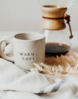 Cream and brown stoneware mug with black text 'warm + cozy' on the front, in front of coffee cafetiere, on striped tea towel.