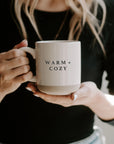 Cream and brown stoneware mug with black text 'warm + cozy' on the front held up with handle and underneath.