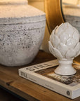 White ceramic artichoke on stand, styled with white decorative book and lamp.