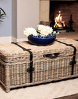 Wicker basket coffee table styled with blue bobble bowl with flowers in, in front of fire on white rug.