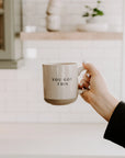 Cream and brown stoneware mug with black 'you got this' text embossed, held up by the handle.