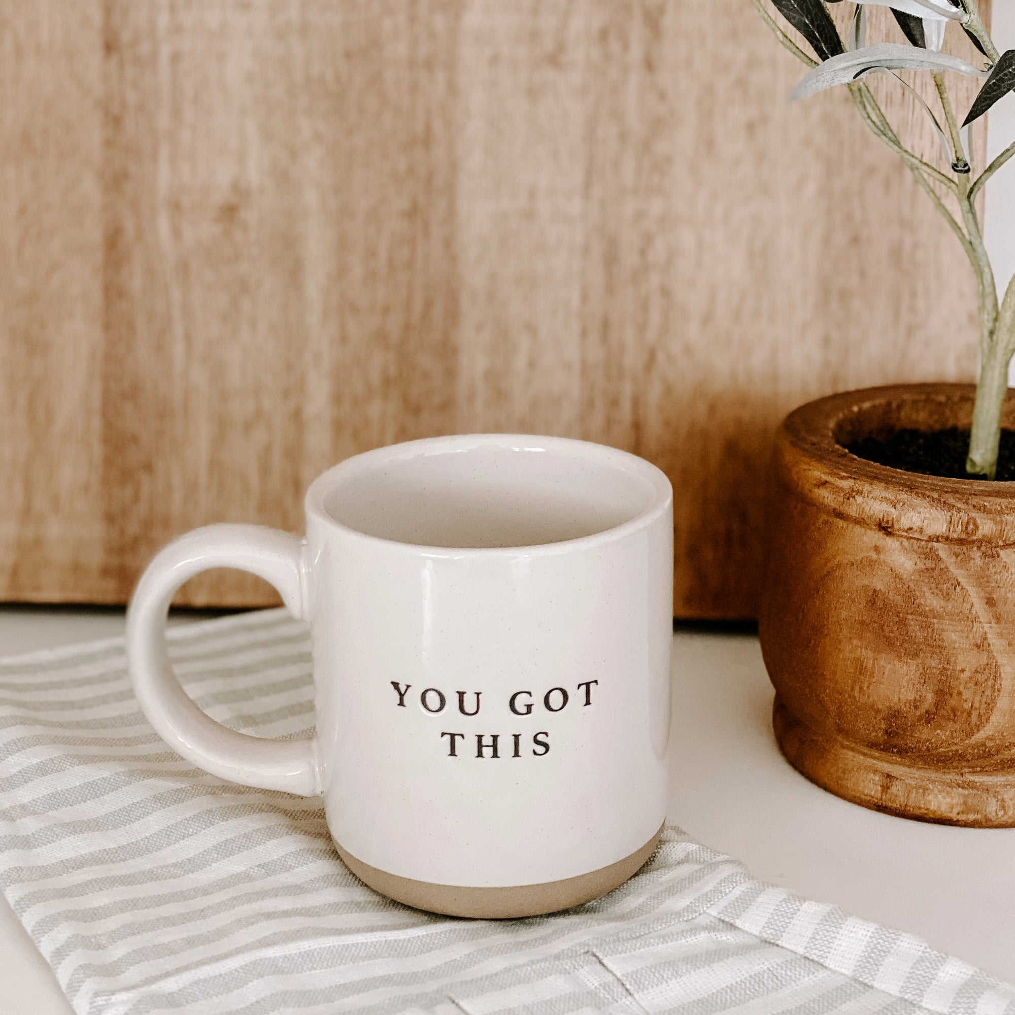 Cream and brown stoneware mug with black 'you got this' text embossed on a striped tea towel in front of plant.