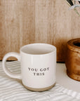 Cream and brown stoneware mug with black 'you got this' text embossed on a striped tea towel in front of plant.