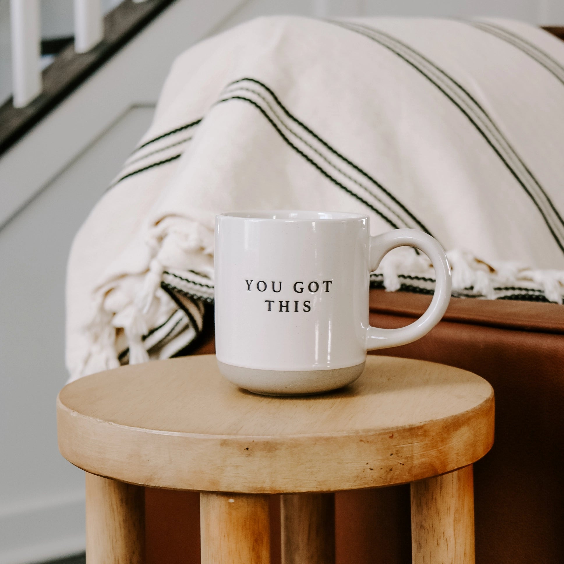 Cream and brown stoneware mug with black 'you got this' text embossed, on a wooden side table in front of sofa.