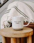 Cream and brown stoneware mug with black 'you got this' text embossed, on a wooden side table in front of sofa.