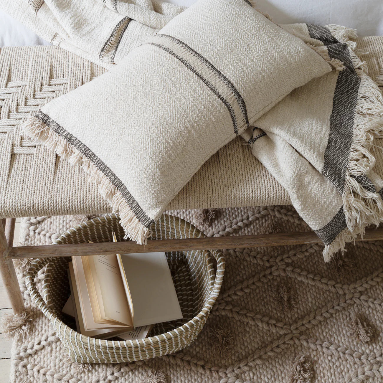 Textured cotton throw blanket and matching cushion on a woven bench with a chunky rug beneath.