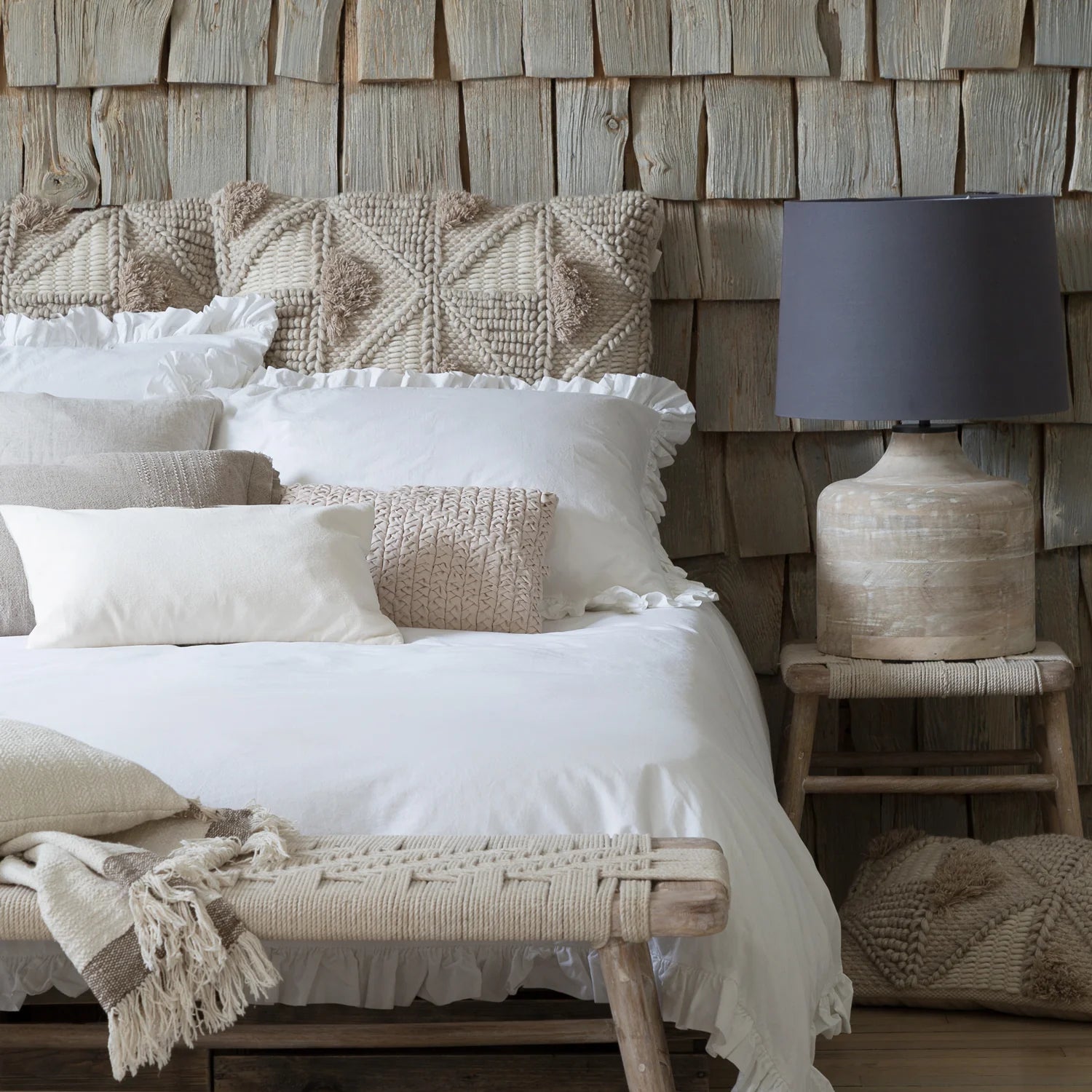 a rustic bedroom with wooden cladded wall and white frilly bedding, the woven bench is at the foot of the bed,